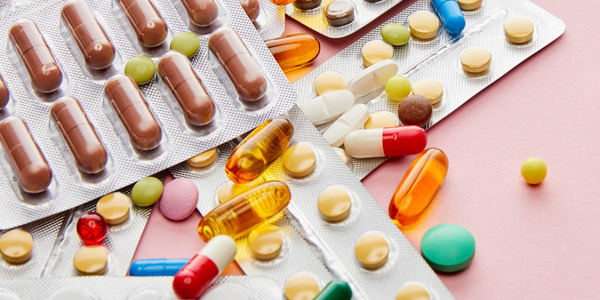 Mehrere Blisterpackungen mit Tabletten und Kapseln in verschiedenen Farben, zusätzlich lose Pillen auf einer rosa Oberfläche verteilt.